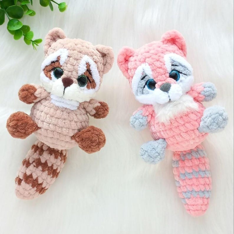 May include: Two crocheted raccoon plush toys. One is tan and brown, the other is pink and gray. Both have white accents around the eyes and muzzle, with black eyes. The raccoons have small arms and legs and a striped tail.
