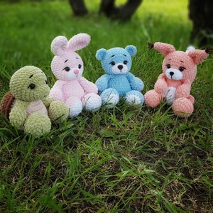 May include: Four crocheted stuffed toys: a pink bunny, a blue bear, a green turtle, and a pink fox. The toys are sitting on a bed of green grass.