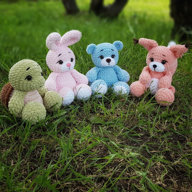 May include: Four crocheted stuffed toys: a pink bunny, a blue bear, a green turtle, and a pink fox. The toys are sitting on a bed of green grass.