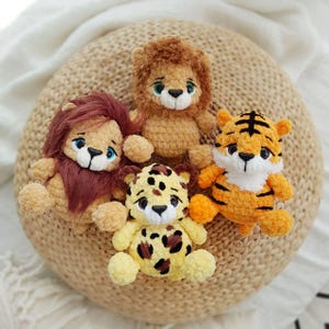 May include: Four crocheted stuffed animals: a lion, a tiger, a leopard, and a lion with a brown mane. The animals are sitting on a woven straw mat.