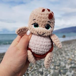 May include: A crocheted turtle plushie in shades of beige, brown, and light pink. The turtle has a smiling face with black eyes and green highlights. The plush toy is held in front of a blurred beach background.