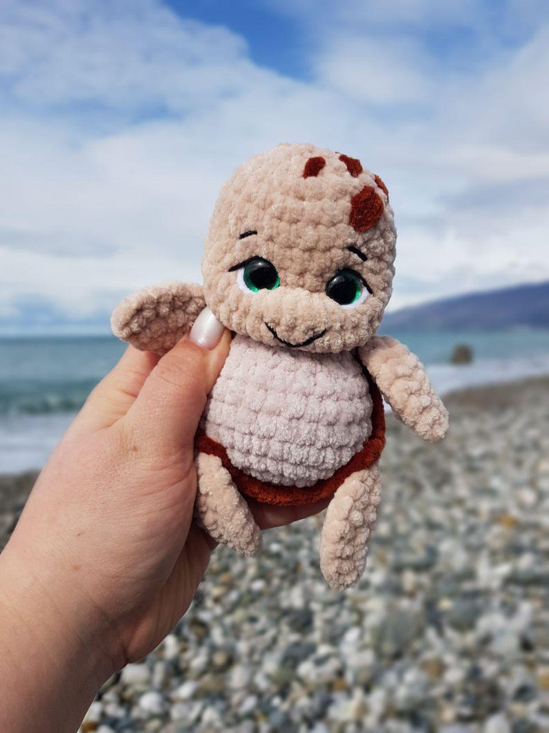 May include: A crocheted turtle plushie in shades of beige, brown, and light pink. The turtle has a smiling face with black eyes and green highlights. The plush toy is held in front of a blurred beach background.