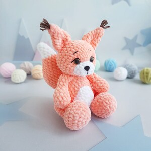 May include: A crocheted plush toy in the shape of a fox with a peach coloured body and white accents. The fox has a long, bushy tail and a sweet expression.