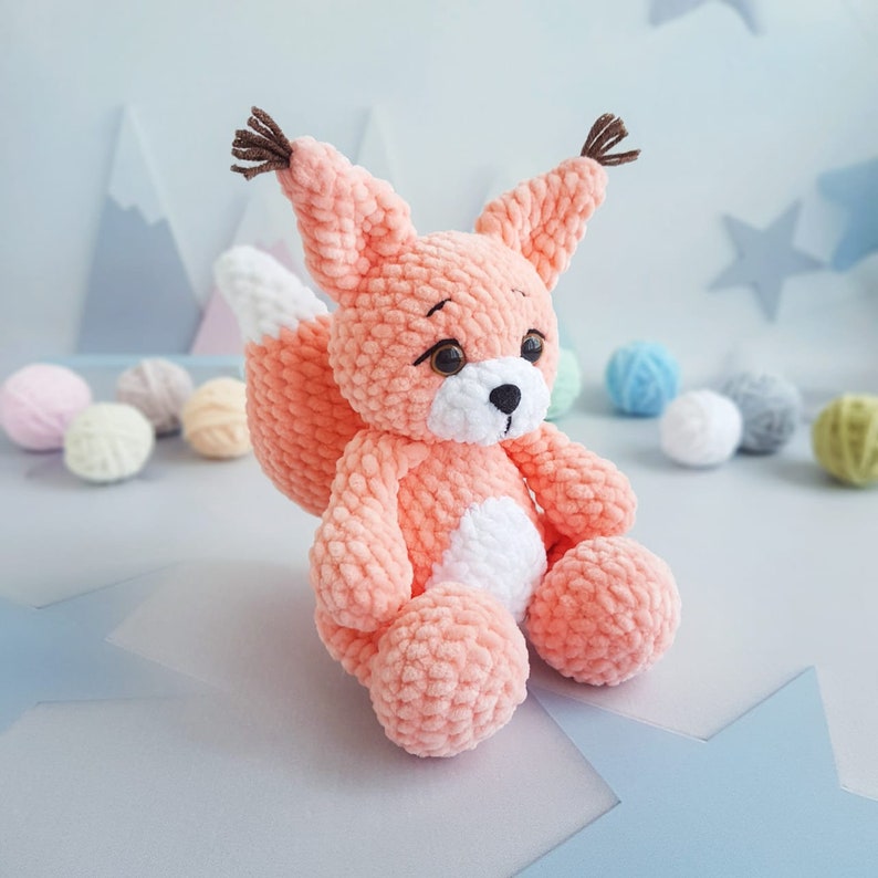May include: A crocheted plush toy in the shape of a fox with a peach coloured body and white accents. The fox has a long, bushy tail and a sweet expression.