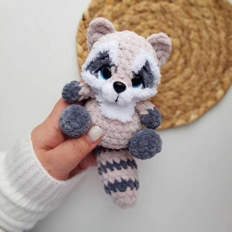 May include: A handmade crochet raccoon plush toy. The raccoon has a light brown body, white accents, and a gray tail, paws, and ears. It has blue eyes and a white muzzle. The plush is held in a hand.