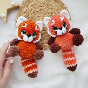 May include: Two handmade crochet red panda stuffed animals. The plush toys have orange bodies, brown limbs, and white accents on their faces and ears. They have black eyes and striped tails. The toys are displayed on a white surface.
