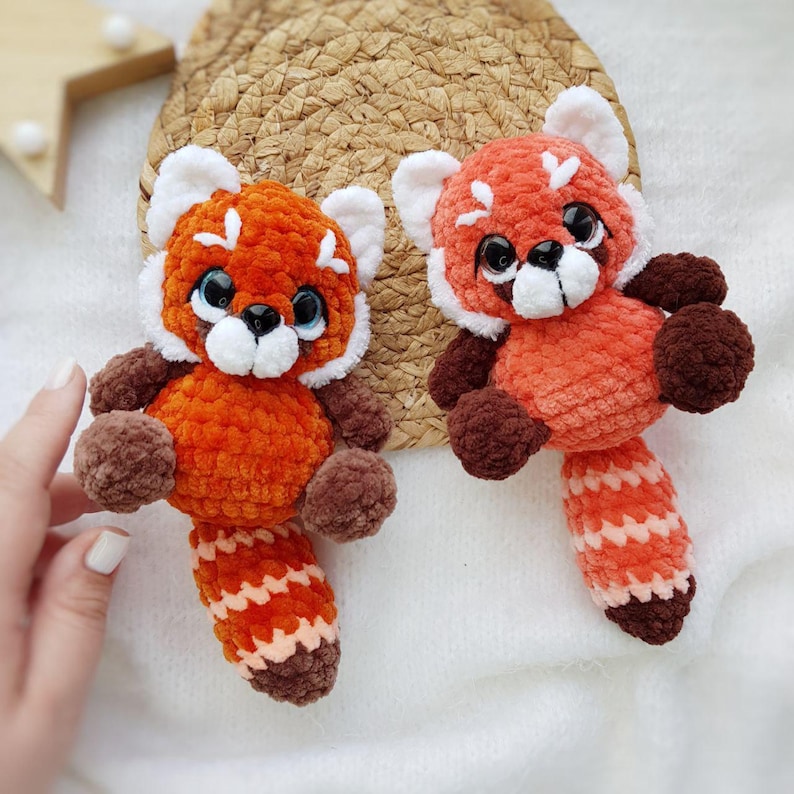 May include: Two handmade crochet red panda stuffed animals. The plush toys have orange bodies, brown limbs, and white accents on their faces and ears. They have black eyes and striped tails. The toys are displayed on a white surface.