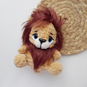 May include: A crocheted lion plush toy with brown fur, a white face, and blue eyes. The lion is sitting on a white surface.
