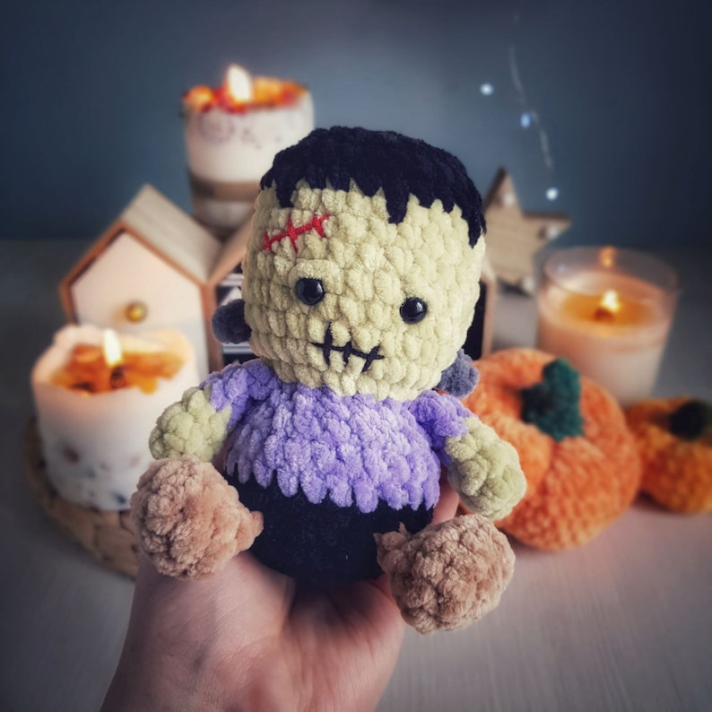 May include: A crocheted plush toy in the shape of a monster with a purple shirt, black pants, and a stitched-on red scar. The toy is sitting on a hand with a blurry background of pumpkins and candles.