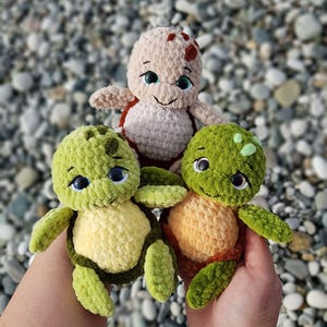May include: Three handmade crochet turtle plushies in shades of green, tan, and orange. The turtles have smiling faces, big eyes, and are held in a person's hands. The background is a blurred view of pebbles.