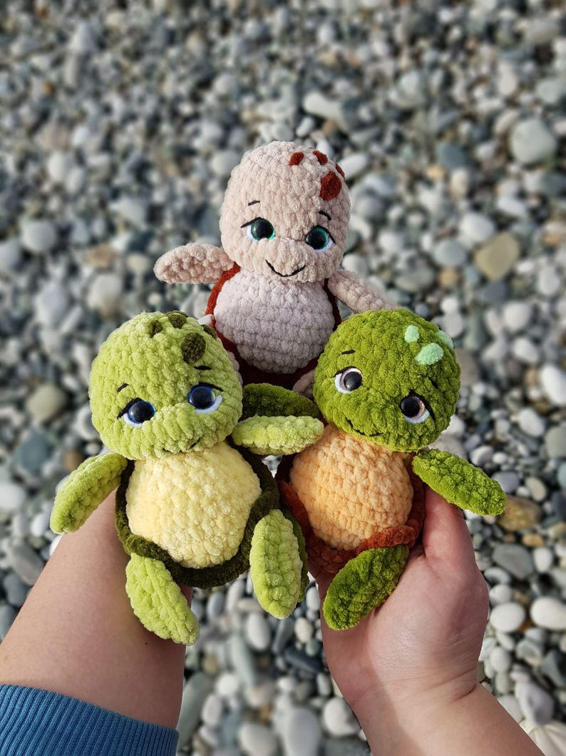 May include: Three handmade crochet turtle plushies in shades of green, tan, and orange. The turtles have smiling faces, big eyes, and are held in a person's hands. The background is a blurred view of pebbles.