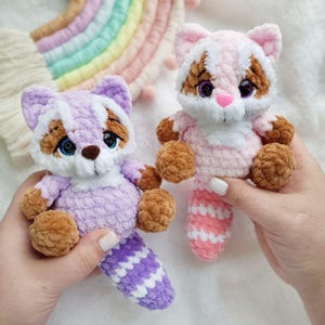 May include: Two handmade crochet raccoon plush toys. One is lavender with a white face and brown accents, the other is pink with brown accents. Both have brown paws and striped tails. The toys are held up by hands.