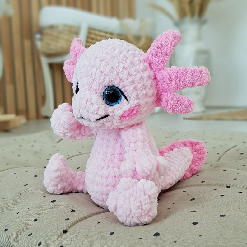 Pode incluir: Um brinquedo de pel&uacute;cia axolotl de croch&ecirc; rosa com olhos azuis. O brinquedo est&aacute; sentado em uma superf&iacute;cie com padr&atilde;o marrom e branco.