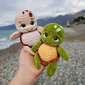 May include: Two handmade crochet turtle plushies. One is tan with brown accents, the other is green and orange. Both have large, expressive eyes and are held in a hand against a blurred beach background.