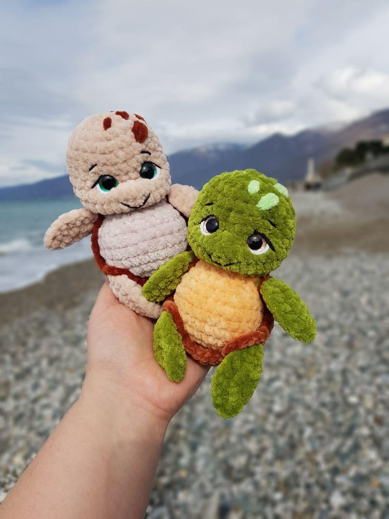 May include: Two handmade crochet turtle plushies. One is tan with brown accents, the other is green and orange. Both have large, expressive eyes and are held in a hand against a blurred beach background.