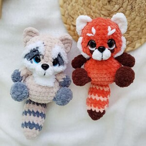 May include: Two handmade crochet animal plushies: a raccoon and a red panda. The raccoon is beige, gray, and blue, with a striped tail. The red panda is orange, brown, and white, with a striped tail. Both have black eyes.