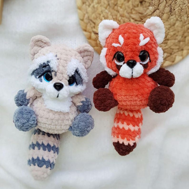 May include: Two handmade crochet animal plushies: a raccoon and a red panda. The raccoon is beige, gray, and blue, with a striped tail. The red panda is orange, brown, and white, with a striped tail. Both have black eyes.
