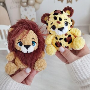 May include: Two crocheted stuffed animals, a lion and a cheetah, are held in the hands of a person. The lion is tan with a brown mane and blue eyes. The cheetah is yellow with brown spots and blue eyes.
