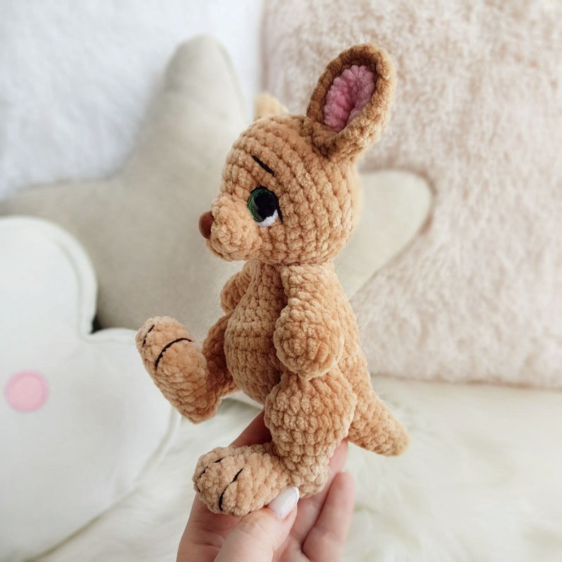 May include: A handcrafted, tan crochet kangaroo toy. The kangaroo features large green eyes, a brown nose, and pink inner ears. The plush toy is approximately 15 cm tall and is posed sitting down. The soft toy is made with a detailed crochet design.