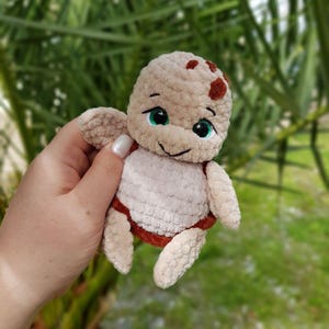 May include: A handmade crochet turtle plushie in beige, brown, and white. The turtle has a smiling face with green eyes and small flipper-like arms and legs. The plushie is held against a blurred green background.