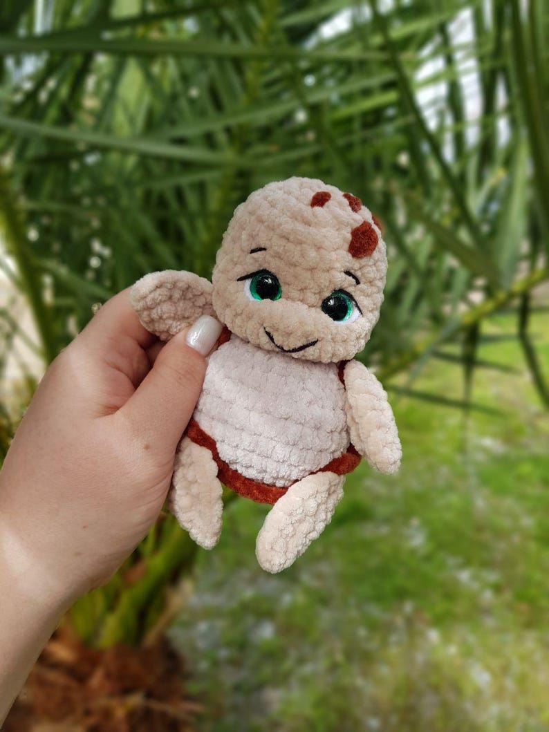 May include: A handmade crochet turtle plushie in beige, brown, and white. The turtle has a smiling face with green eyes and small flipper-like arms and legs. The plushie is held against a blurred green background.
