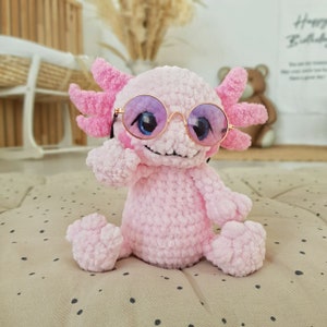 Pode incluir: Um brinquedo de pel&uacute;cia axolotl de croch&ecirc; rosa usando &oacute;culos de sol rosa e roxo. O axolotl est&aacute; sentado em uma superf&iacute;cie de bolinhas brancas e marrons.