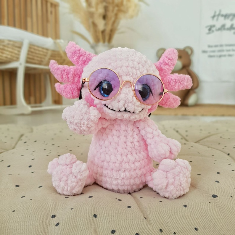 Pode incluir: Um brinquedo de pel&uacute;cia axolotl de croch&ecirc; rosa usando &oacute;culos de sol rosa e roxo. O axolotl est&aacute; sentado em uma superf&iacute;cie de bolinhas brancas e marrons.