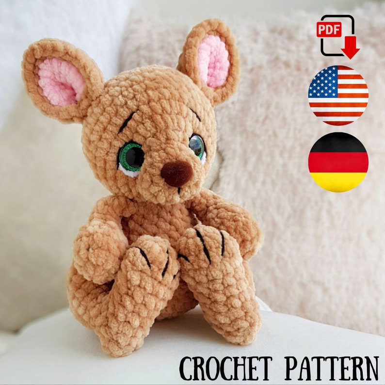 May include: A crocheted brown mouse toy with pink inner ears, green eyes, and a brown nose. The mouse is sitting with its paws forward. The image includes the text "CROCHET PATTERN" and a PDF download icon with the American flag.