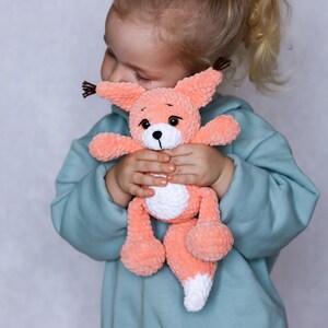 May include: A young child is holding a crocheted plush toy fox in their arms. The fox is peach coloured with white accents and brown eyes. The child is wearing a light blue sweatshirt.