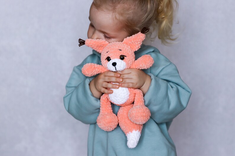 May include: A young child is holding a crocheted plush toy fox in their arms. The fox is peach coloured with white accents and brown eyes. The child is wearing a light blue sweatshirt.
