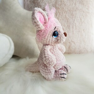 May include: A light pink crochet kangaroo toy with a pink lace headband. The kangaroo has blue eyes, a brown nose, and black claws. The plush is sitting on a white, fluffy surface.