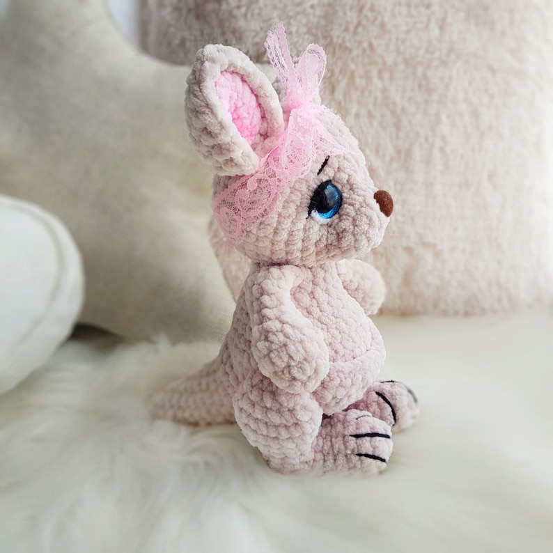 May include: A light pink crochet kangaroo toy with a pink lace headband. The kangaroo has blue eyes, a brown nose, and black claws. The plush is sitting on a white, fluffy surface.