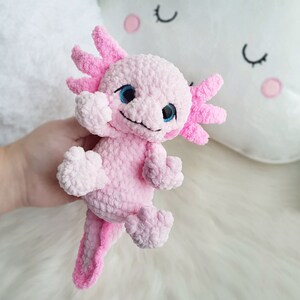 Pode incluir: Um brinquedo de pel&uacute;cia axolotl de croch&ecirc; rosa com olhos azuis. O brinquedo est&aacute; sentado em uma superf&iacute;cie branca e fofa.