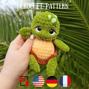 May include: A handmade crochet turtle plushie with a green head and limbs, a tan shell, and brown accents. The text "CROCHET PATTERN" is at the top. The plushie is held in a hand. Flags of the USA, Germany, and France are at the bottom.