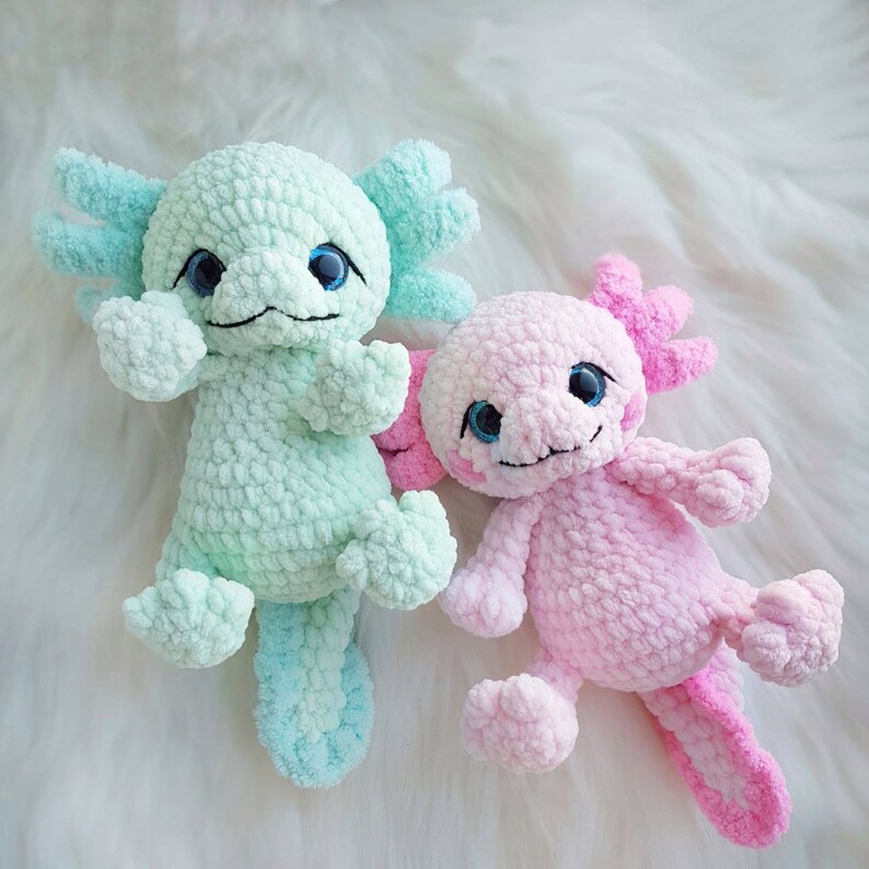 Pode incluir: Dois peluches de axolotl de croch&ecirc;, um na cor verde claro e o outro na cor rosa claro. Ambos os peluches t&ecirc;m olhos grandes e uma express&atilde;o sorridente.