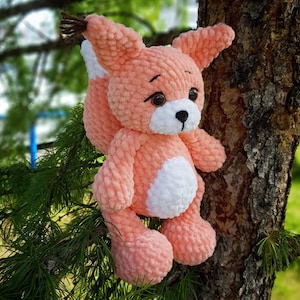 May include: A crocheted plush fox toy with a pink body, white belly, and brown eyes. The toy is sitting on a tree branch.