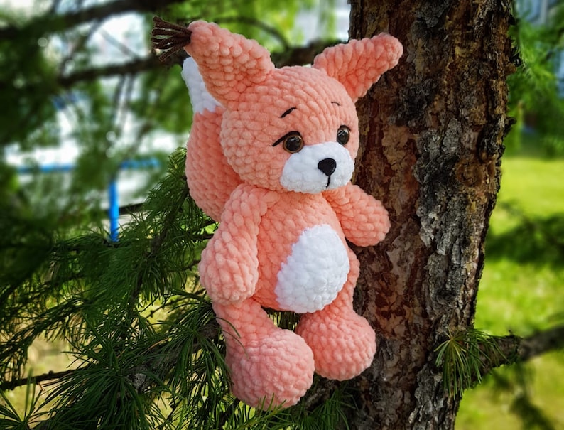 May include: A crocheted plush fox toy with a pink body, white belly, and brown eyes. The toy is sitting on a tree branch.