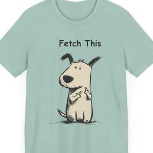 Może przedstawiać: Jasnozielona koszulka z ilustracją psa z kreskówki. Pies jest beżowy z czarnymi akcentami i pokazuje środkowy palec. Tekst "Fetch This" znajduje się nad psem.