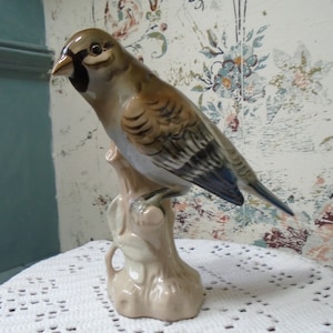 Op de afbeelding: Een porseleinen beeldje van een vogel die op een tak zit. De vogel is geschilderd in tinten bruin, grijs en blauw. Het beeldje is een vintage stuk en heeft een delicaat, handgeschilderd ontwerp.