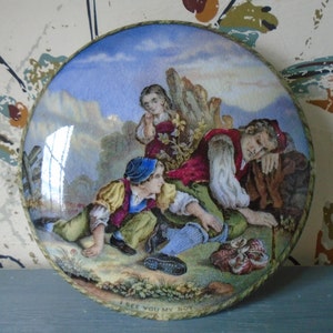 Könnte beinhalten: Ein runder Keramikdeckel mit einer farbenfrohen bemalten Szene von drei Figuren in einer Landschaft. Die Figuren sind ein Mann, eine Frau und ein Kind. Der Mann trägt ein rot-blaues Outfit und liegt auf dem Boden. Die Frau trägt ein blau-weißes Kleid und steht. Das Kind trägt ein gelb-rotes Outfit und sitzt. Die Szene spielt auf einem Feld mit Bäumen und blauem Himmel. Der Deckel hat einen goldenen Rand und die Worte "I SEE YOU MY BOY" sind unten in Schwarz geschrieben.