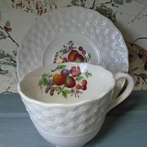 May include: White porcelain teacup and saucer with a raised floral design. The teacup features a painted design of fruit and flowers in shades of red, yellow, green, and purple.