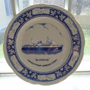 May include: A white ceramic plate with a blue and white design featuring a ship called "Bluenose" and the text "Yarmouth-Bar Harbour Ferry".