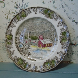 May include: A vintage ceramic plate with a winter scene featuring a red schoolhouse, snow-covered trees, and a snowy landscape. The plate has a green and brown floral border.