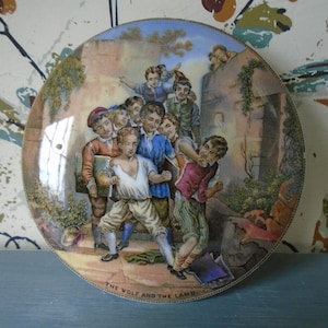 May include: A vintage ceramic plate lid featuring a colourful illustration of children at play. The scene shows a group of children, some holding books, with the text "THE WOLF AND THE LAMB." printed below the image.