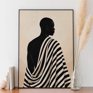 May include: A framed art print featuring a silhouette of a person draped in a black and white striped fabric. The figure is set against a beige background. The artwork is displayed on a wooden surface.