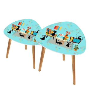 May include: Two retro-style side tables with a light blue surface featuring a mid-century modern living room scene with black cats. The tables have wooden legs and a unique, rounded triangular shape.