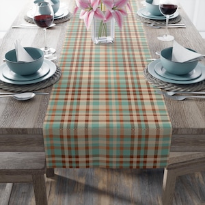 Op de afbeelding: Een geruite tafelloper in tinten van teal, bruin en beige, gepresenteerd op een houten tafel. De loper heeft een klassiek ruitpatroon. De tafel is gedekt met teal kommen, witte borden, bestek en wijnglazen, met een vaas met roze bloemen.
