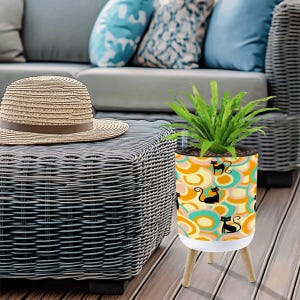 May include: A decorative planter with a retro design featuring black cat silhouettes on a pattern of orange, yellow, and teal swirls. The planter sits on three wooden legs and holds a green plant. A straw hat rests on a nearby wicker table.