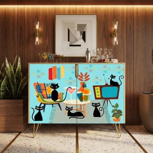May include: A rectangular cabinet with light blue doors featuring a colorful graphic print of four black cats, a mid-century armchair, a television set, a cocktail glass, and a vase. The cabinet is supported by four thin, gold-toned metal legs.