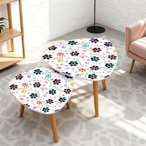 May include: Two white, triangular side tables with wooden legs. The tabletop features a repeating pattern of colorful paw prints in black, red, yellow, blue, and purple. The tables are different sizes and are placed one in front of the other.